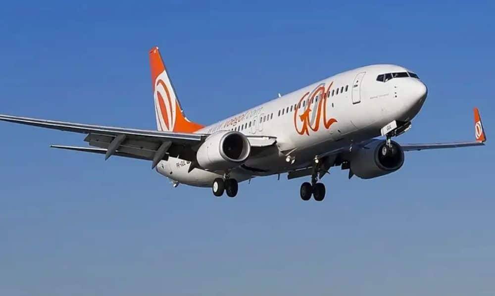 Avião da companhia aérea GOL Linhas Aéreas voando no céu azul claro, em ângulo inferior mostrando o trem de pouso estendido.