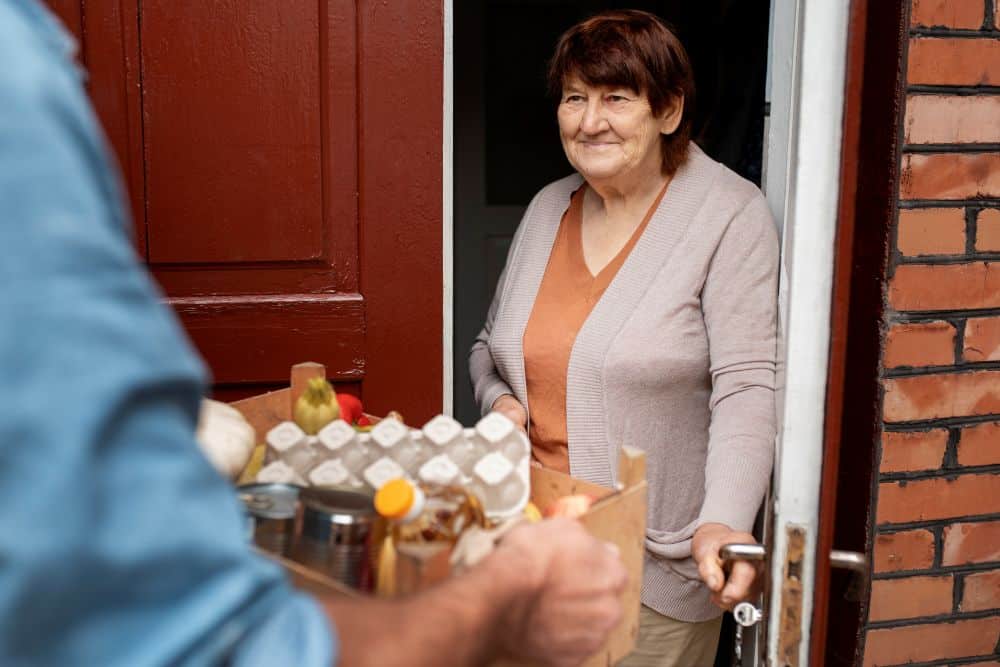 Uma mulher idosa sorridente na porta de sua casa recebe uma caixa de mantimentos de um homem que está de costas, em primeiro plano.