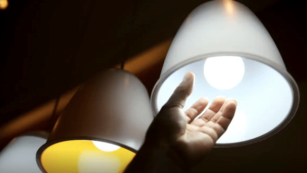 Uma mão alcançando uma lâmpada de LED acesa dentro de uma luminária branca em formato de cone. Ao fundo, outras duas luminárias similares aparecem desfocadas, uma delas com luz amarelada.