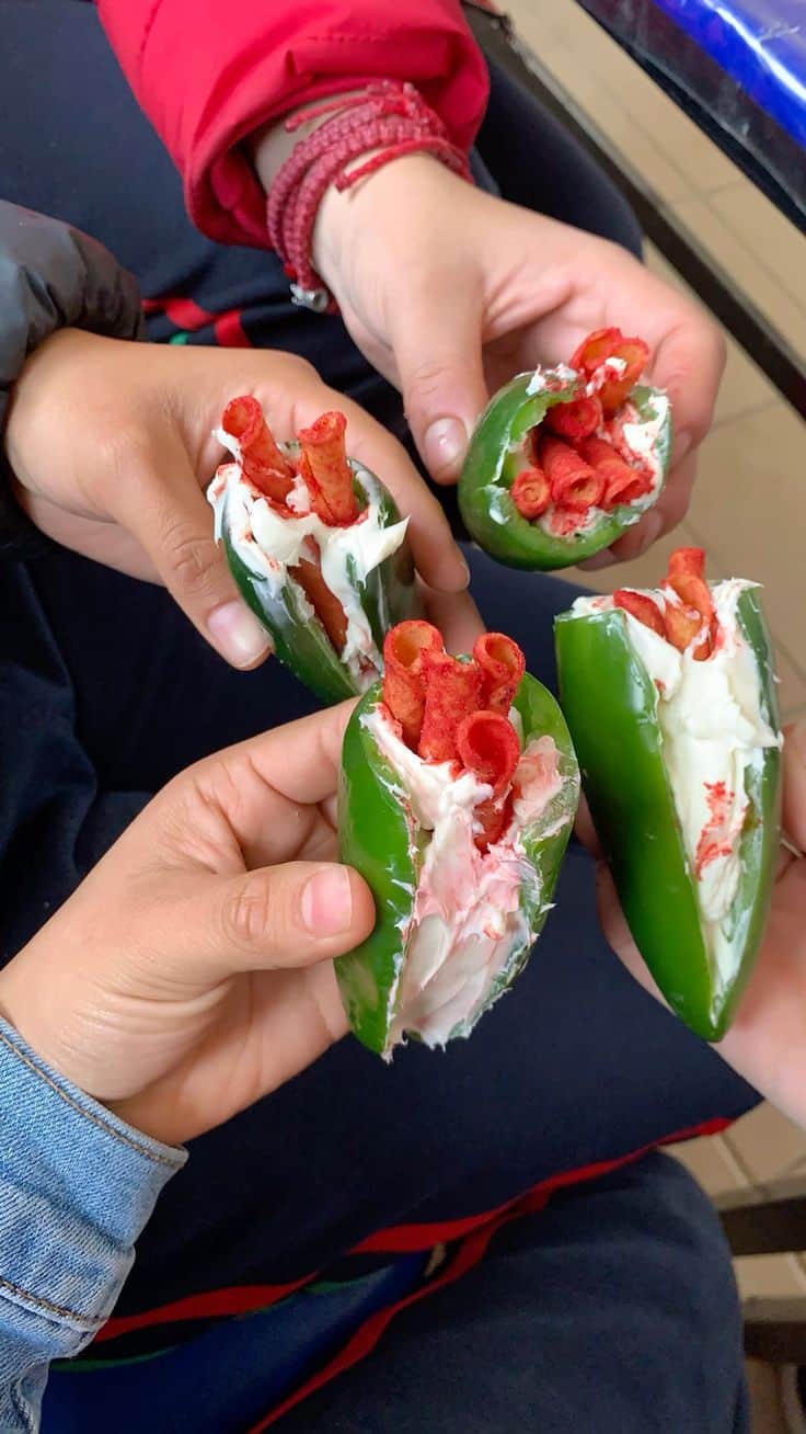 Três pares de mãos segurando quatro pedaços de pimentão verde. Cada pimentão está cortado ao meio e recheado com cream cheese branco, pronto para a cobertura. Vários snacks vermelhos e cilíndricos, Takis Fuego, estão visíveis perto do recheio. Os fundos das roupas são uma jaqueta vermelha, uma camisa azul-marinho e uma manga jeans.