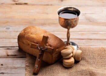 Elementos religiosos sobre mesa de madeira: um pão rústico, um cálice de metal com vinho tinto, um pequeno crucifixo de madeira e pães ázimos sobre um tecido de juta, simbolizando a Santa Ceia e a Sexta-feira Santa.