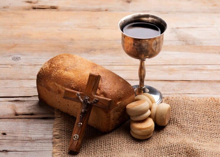 Elementos religiosos sobre mesa de madeira: um pão rústico, um cálice de metal com vinho tinto, um pequeno crucifixo de madeira e pães ázimos sobre um tecido de juta, simbolizando a Santa Ceia e a Sexta-feira Santa.
