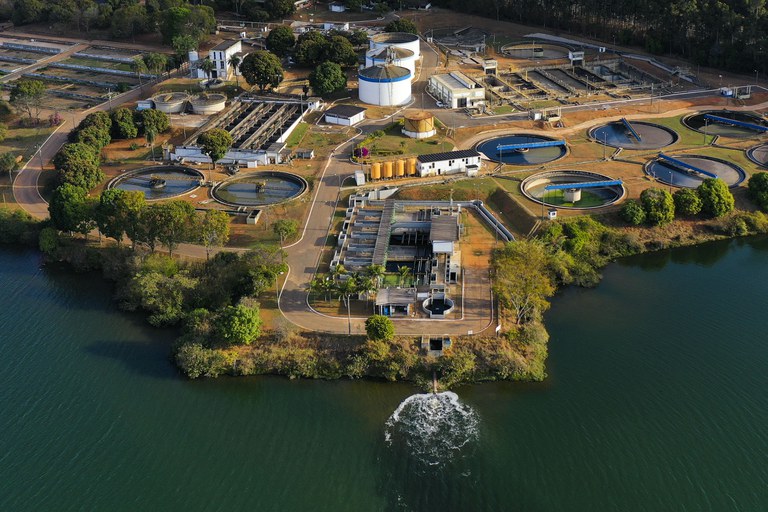Vista aérea de uma estação de tratamento de água e esgoto localizada à beira de um rio ou represa. A imagem mostra tanques circulares de decantação, reservatórios cilíndricos e estruturas de filtragem cercadas por árvores e vias internas.