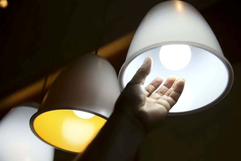 Mão estendida em direção a uma luminária branca suspensa acesa, de onde emana uma luz branca brilhante. Ao lado, outras luminárias semelhantes estão visíveis, sendo uma delas de cor amarela por dentro e acesa. O fundo é escuro.