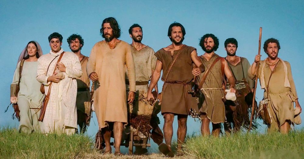 Grupo de atores caracterizados como Jesus e seus discípulos caminhando juntos em um campo sob o céu azul.