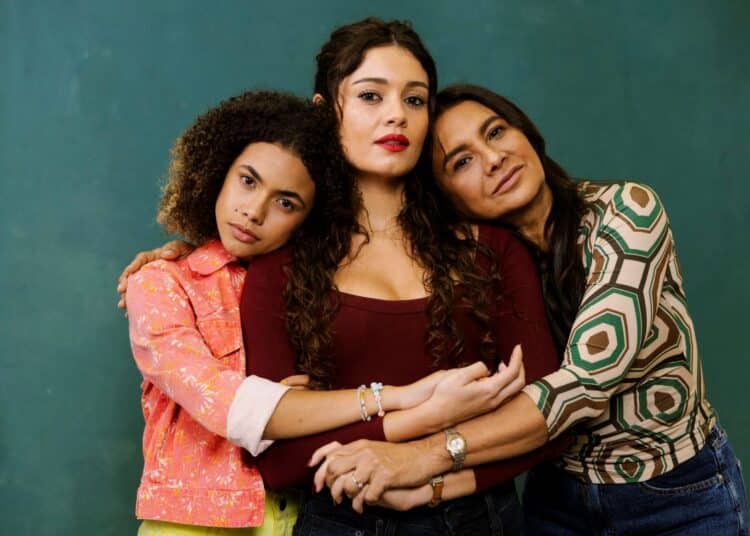 Alana Cabral, Sophie Charlotte e Dira Paes posam juntas em um retrato promocional. Alana Cabral, à esquerda com cabelo cacheado e jaqueta estampada, abraça Sophie Charlotte (centro), que veste blusa bordô. Dira Paes, à direita com blusa geométrica, apoia a cabeça em Sophie Charlotte, com as mãos de Alana e Dira cruzadas sobre as de Sophie. O fundo é verde escuro.