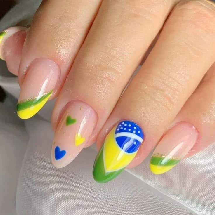 Close-up de unhas em formato amendoado com base nude translúcida. A decoração é variada: uma unha traz o desenho da bandeira do Brasil com detalhes em azul e branco; outra apresenta três pequenos corações nas cores verde, amarelo e azul; e as demais possuem uma francesinha dupla curvada em verde e amarelo com um traço fino de glitter dourado. A mão repousa sobre um tecido branco.