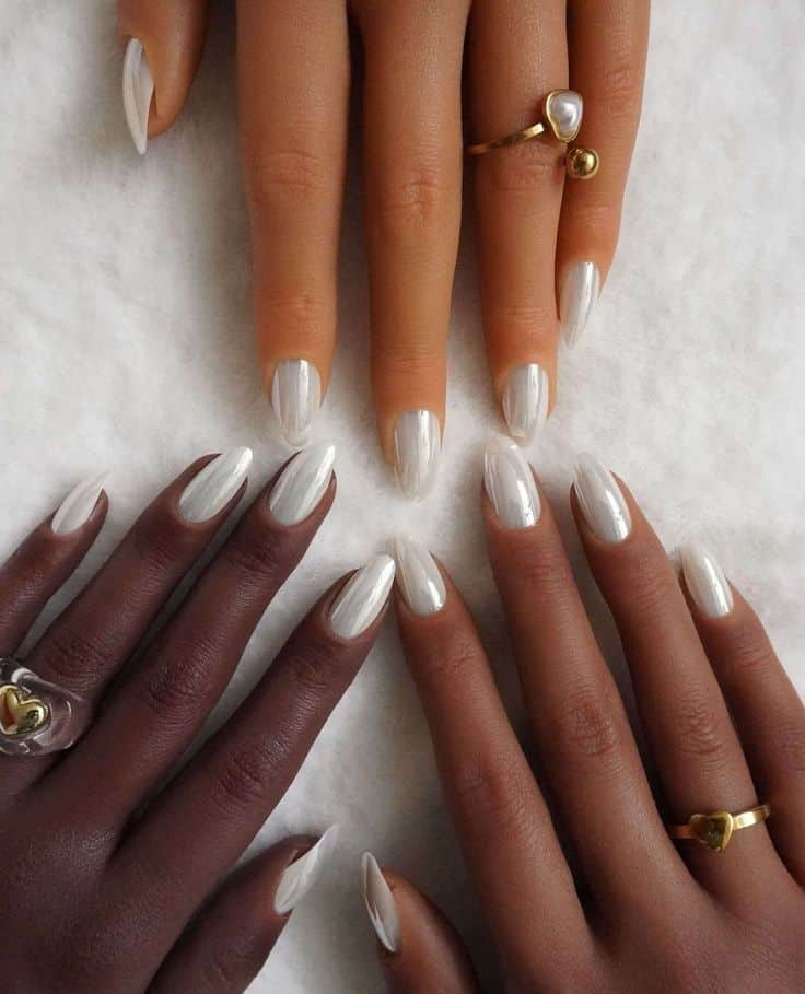 Close de três mãos com diferentes tons de pele (clara, morena e negra) exibindo unhas longas em formato amendoado com efeito chrome white espelhado sobre um fundo felpudo branco. As mãos usam anéis dourados delicados.
