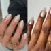 Duas mãos femininas lado a lado: a da esquerda tem pele clara e anéis dourados, com unhas amendoadas pintadas de branco cintilante iridescente. A da direita tem pele negra e um anel de laço dourado, com unhas longas de branco cintilante opaco. Ambas sobre fundo neutro.