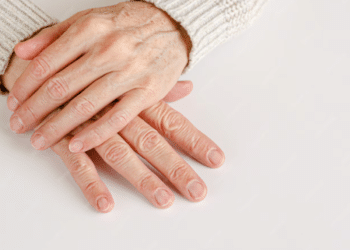Duas mãos maduras entrelaçadas sobre uma superfície branca, mostrando unhas naturais, limpas e bem cuidadas após os 60 anos.