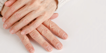 Duas mãos maduras entrelaçadas sobre uma superfície branca, mostrando unhas naturais, limpas e bem cuidadas após os 60 anos.