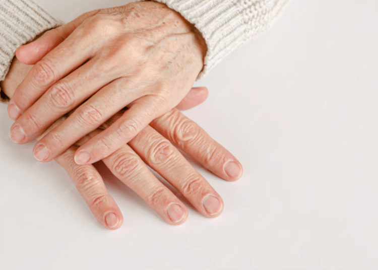 Duas mãos maduras entrelaçadas sobre uma superfície branca, mostrando unhas naturais, limpas e bem cuidadas após os 60 anos.