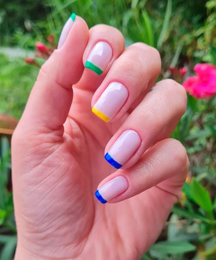 Close-up da mão de uma pessoa com as unhas de formato quadrado, pintadas com uma base rosa nude translúcida. As pontas das unhas possuem uma francesinha colorida e vibrante: a unha do dedo indicador é verde, a do dedo médio é amarela, e a do dedo anelar e do mindinho são azuis. A mão está posicionada sobre um fundo de folhagem verde e flores rosa embaçadas, em um ambiente externo com luz natural.