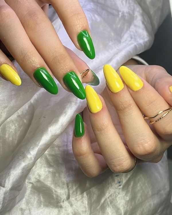 Close-up das mãos de uma pessoa com unhas longas em formato amendoado, pintadas intercalando as cores verde bandeira e amarelo vibrante com acabamento brilhante. A pessoa usa anéis dourados finos e delicados, e as mãos estão posicionadas sobre um tecido branco acetinado.