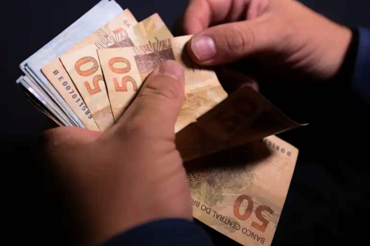Mãos de uma pessoa contando notas de 50 reais brasileiros sobre um fundo escuro.