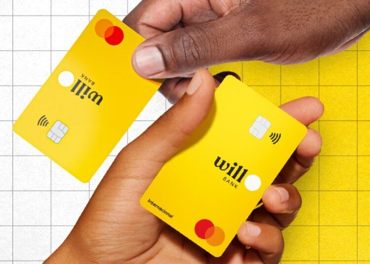 Duas mãos de tons de pele diferentes (uma mais escura e uma mais clara) segurando e passando entre si dois cartões de crédito e débito Will Bank. Os cartões são de cor amarela vibrante e laranjas, com o logotipo "WILL BANK" em preto e o símbolo da Mastercard em vermelho e laranja visíveis. O fundo é dividido entre um padrão de malha de grade branca e uma textura de concreto amarela.