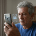 Homem idoso de cabelos grisalhos segura smartphone com ícone de reconhecimento facial, usando suéter azul em ambiente residencial