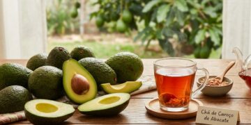 Xícara de vidro com chá de caroço de abacate fumegante ao lado de abacates inteiros e cortados sobre mesa de madeira