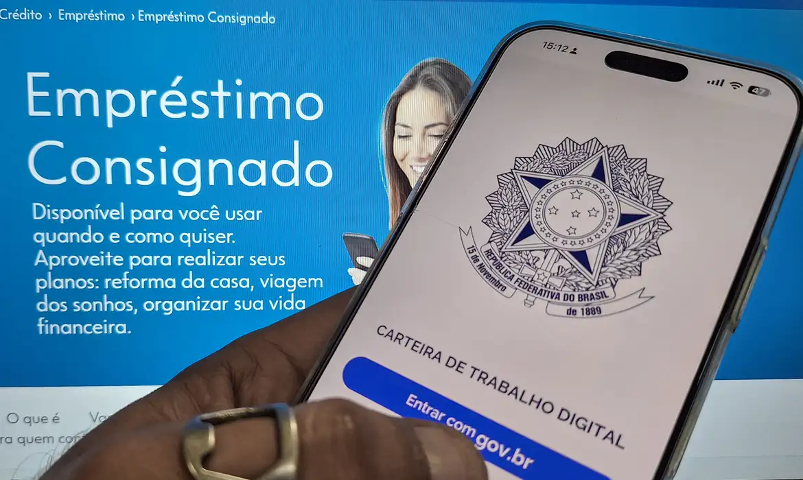 Mão segura smartphone com tela da Carteira de Trabalho Digital diante de site sobre empréstimo consignado