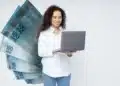 Mulher de cabelos cacheados usando camisa branca segura laptop cinza com notas de 100 reais em leque ao lado
