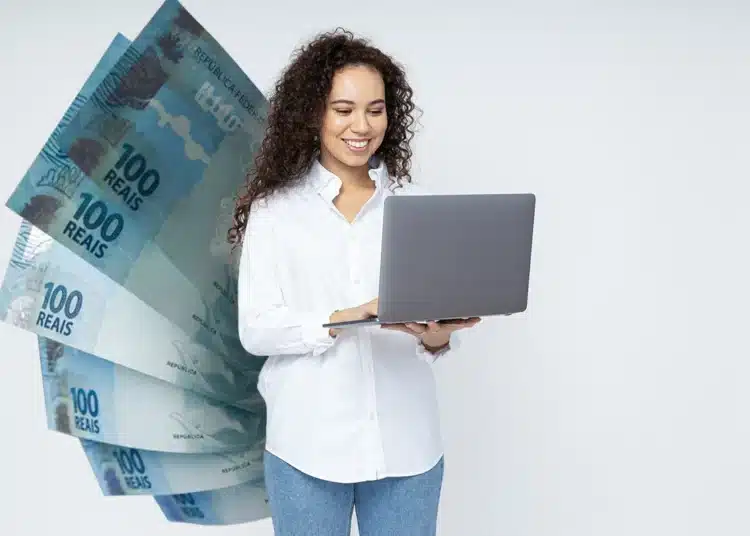 Mulher de cabelos cacheados usando camisa branca segura laptop cinza com notas de 100 reais em leque ao lado