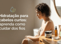 Mulher de costas, cabelos cacheados curtos castanhos, usa toalha bege em frente ao espelho com produtos capilares na bancada