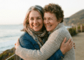 Duas mulheres sorrindo se abraçam em trilha costeira com mar e montanha ao fundo durante pôr do sol