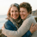 Duas mulheres sorrindo se abraçam em trilha costeira com mar e montanha ao fundo durante pôr do sol
