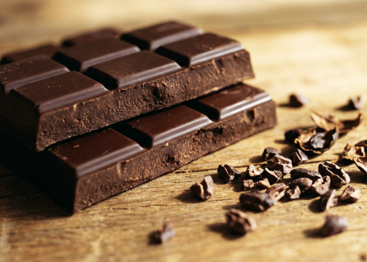 Duas barras de chocolate escuro empilhadas ao lado de nibs de cacau espalhados em superfície de madeira clara