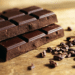 Duas barras de chocolate escuro empilhadas ao lado de nibs de cacau espalhados em superfície de madeira clara