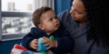 Mulher segura no colo um bebê agasalhado que segura um brinquedo, perto de uma janela