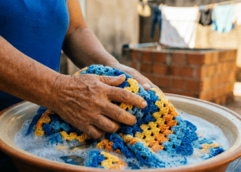 Mãos de mulher idosa lavando manta de crochê em tons de azul, laranja e amarelo em bacia de barro com espuma