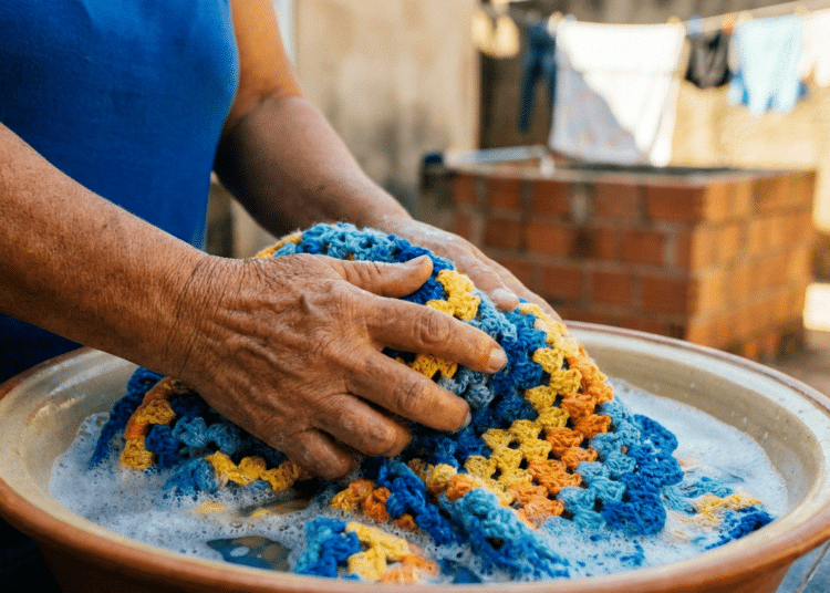 Mãos de mulher idosa lavando manta de crochê em tons de azul, laranja e amarelo em bacia de barro com espuma