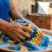 Mãos de mulher idosa lavando manta de crochê em tons de azul, laranja e amarelo em bacia de barro com espuma