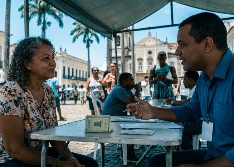Servidor do INSS de camisa azul atende cidadã em mesa sob tenda, com documentos visíveis, no centro histórico de Salvador.