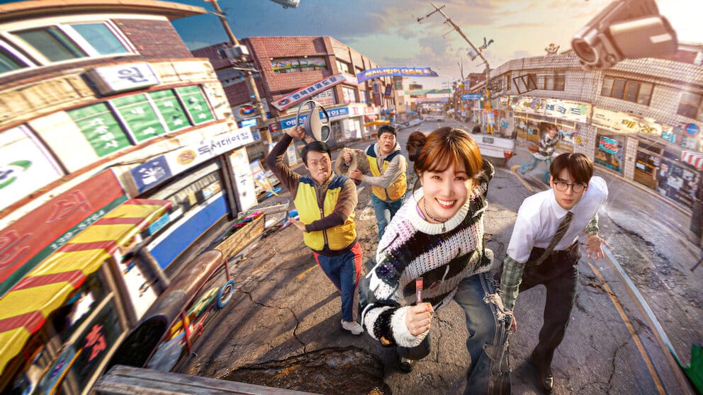Quatro personagens coreanos correndo em direção à câmera em uma rua estreita e inclinada de uma cidade coreana antiga, com prédios coloridos ao fundo. À frente, uma mulher sorridente com blusa listrada e, ao lado, um homem de óculos e camisa social branca.