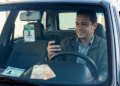 Motorista de app com jaqueta verde e camisa xadrez olha celular no carro, com crachá de identificação e notas de reais no painel.