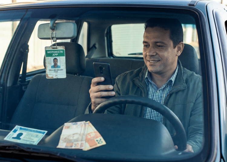 Motorista de app com jaqueta verde e camisa xadrez olha celular no carro, com crachá de identificação e notas de reais no painel.