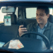 Motorista de app com jaqueta verde e camisa xadrez olha celular no carro, com crachá de identificação e notas de reais no painel.