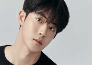 Nam Joo-hyuk, ator coreano de cabelos escuros e camiseta preta, em foto de ensaio com fundo cinza claro