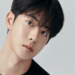 Nam Joo-hyuk, ator coreano de cabelos escuros e camiseta preta, em foto de ensaio com fundo cinza claro