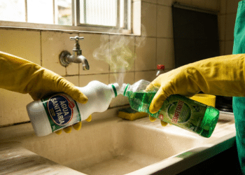 Duas pessoas com luvas amarelas misturam água sanitária e desinfetante, liberando vapor ao lado de uma pia