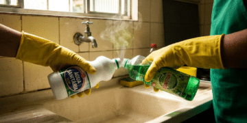 Duas pessoas com luvas amarelas misturam água sanitária e desinfetante, liberando vapor ao lado de uma pia