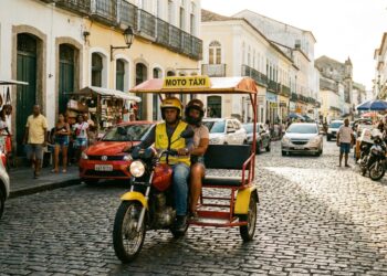 Triciclo moto táxi amarelo com toldo vermelho em rua de paralelepípedos, condutor com colete amarelo e passageiro