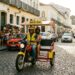 Triciclo moto táxi amarelo com toldo vermelho em rua de paralelepípedos, condutor com colete amarelo e passageiro