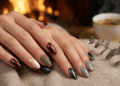 Mãos femininas com unhas amendoadas em esmalte vinho metálico, verde escuro e cinza prateado sobre tecido bege