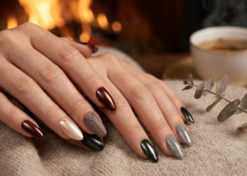 Mãos femininas com unhas amendoadas em esmalte vinho metálico, verde escuro e cinza prateado sobre tecido bege