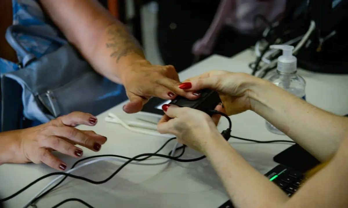 Duas mulheres em mesa, uma coletando digital da outra com leitor eletrônico, frasco de álcool gel ao lado