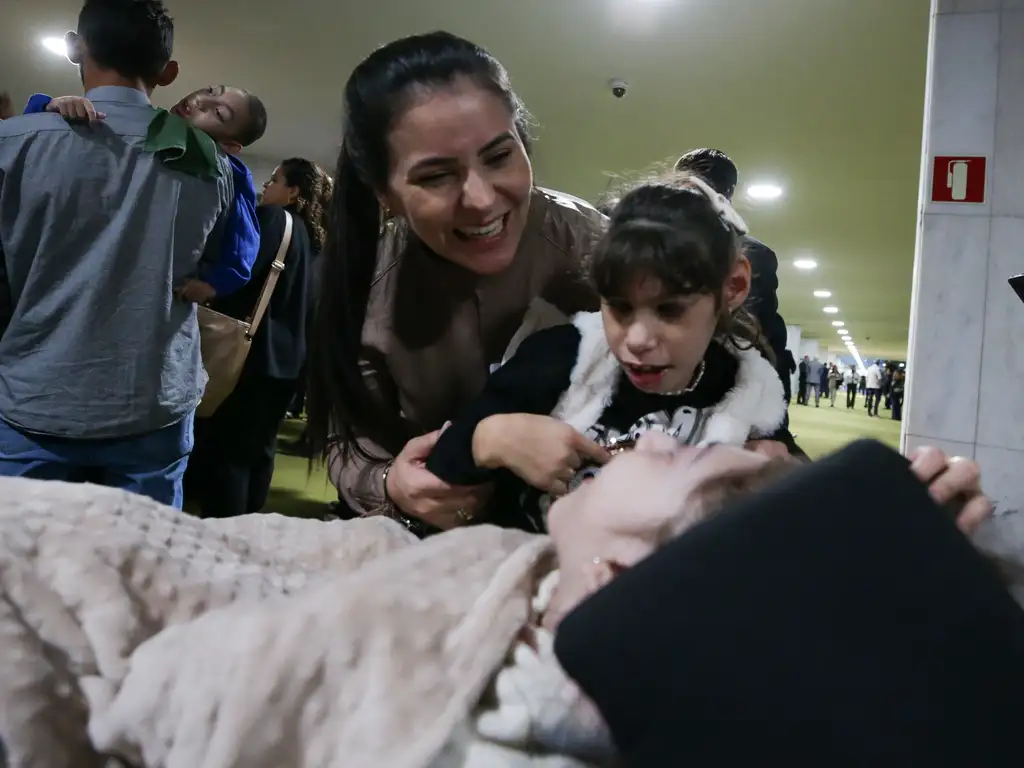 Mulher sorrindo abraça menina ao lado de criança deitada sob cobertor, ambiente de corredores iluminados