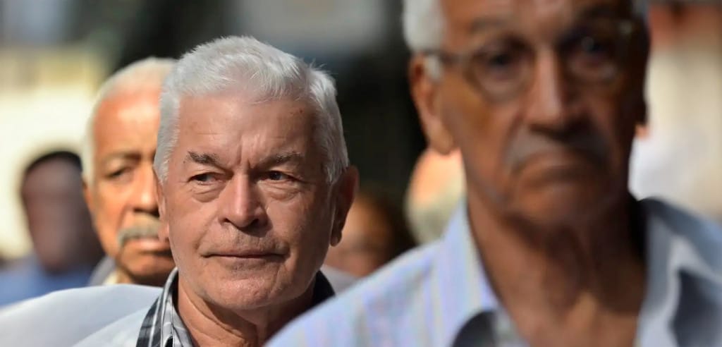 Grupo de homens idosos alinhados em fila, rosto sério, roupas sociais e luz natural